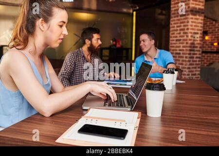 Ernsthafte junge Geschäftsfrau, die an einem großen Tisch im Café sitzt und am Computer arbeitet, wenn ihre Kollegin im Hintergrund Projektdetails diskutiert Stockfoto