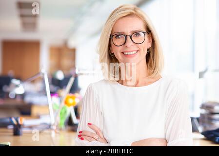 Porträtaufnahme einer attraktiven Geschäftsfrau, die mit im Büro gekreuzten Armen steht, während sie die Kamera betrachtet und lächelt. Stockfoto
