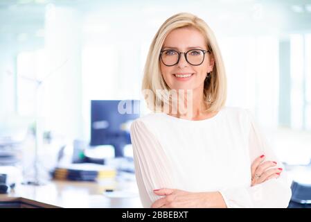 Porträtaufnahme einer attraktiven Geschäftsfrau, die mit im Büro gekreuzten Armen steht, während sie die Kamera betrachtet und lächelt. Stockfoto