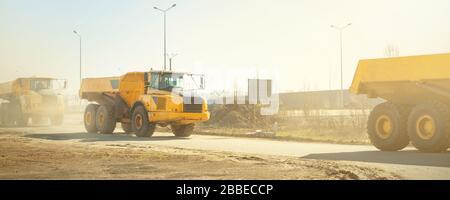 Viele große Gelenkwagen, die am sonnigen Tag mit blauem Himmel auf der neuen Straßenbaustelle fahren, sind mit gelben Müllhalden befahren Stockfoto