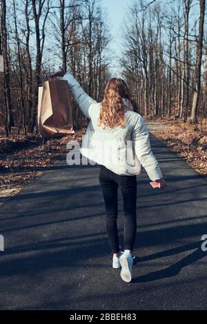 Junge Frau mit großer Papier-Einkaufstasche. Rückansicht des glücklichen Mädchens, das nach dem Einkaufen in einem Park spazieren ging, mit weißer Jacke und Schuhen. Echte Leute, au Stockfoto
