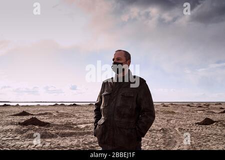 Apokalypse und Kataklysma durch Coronavirus. Reifer Mann in schwarzer Maske und schwarzem Mantel steht mitten in der Wüste, Sands. Einsam, verloren. Surreale Landschaftsap Stockfoto