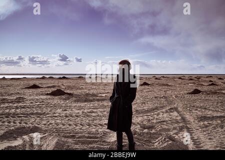 Apokalypse und Kataklysma durch Coronavirus. Frau in schwarzer Maske und schwarzem Mantel steht mitten in der Wüste, Sands. Einsam, verloren. Surreale Landschaft Stockfoto