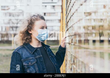 Osteuropäerin besorgt Frau, die im Freien medizinische Maske trägt, neben dem Gitterzaun mit Wohnhaus auf dem Hintergrund steht. Corona-Virus Stockfoto