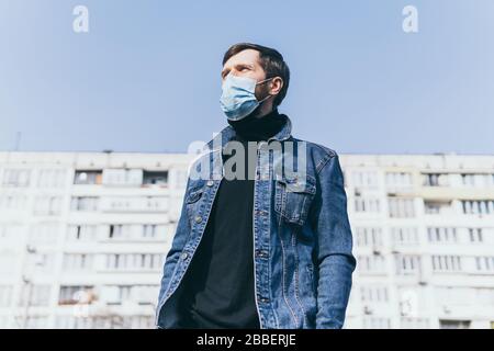 Betroffener junger Mann, der im Freien eine medizinische Maske mit einem Wohnhaus auf dem Hintergrund trägt. Corona-Virus Quarantäne konzeptionelles Bild. Stockfoto