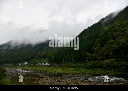 Üppige grüne Landschaft entlang der Takayama-Linie und des Flusses Hida auf dem Weg nach Takayama mit tief hängenden Wolken, die über die Bergketten drapiert sind Stockfoto