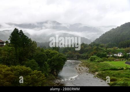 Üppige grüne Landschaft entlang der Takayama-Linie und des Flusses Hida auf dem Weg nach Takayama mit tief hängenden Wolken, die über die Bergketten drapiert sind Stockfoto