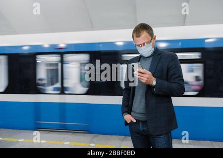 Der männliche Passagier trägt Gesichtsmaskenposen auf der Plattform, wartet auf den Zug, pendelt im Untergrund, konzentriert auf das Smartphone-Gerät, liest online Nachrichten. Viru Stockfoto