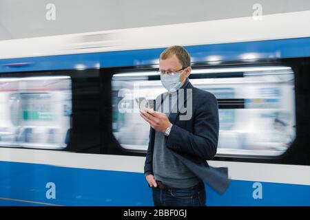 Foto des Mannes in Augenspülung und Schutzmaske, benutzt modernes Telefon, fährt mit öffentlichen Verkehrsmitteln, steht gegen Zug in Bewegung auf Bahnsteig, Gerät Stockfoto