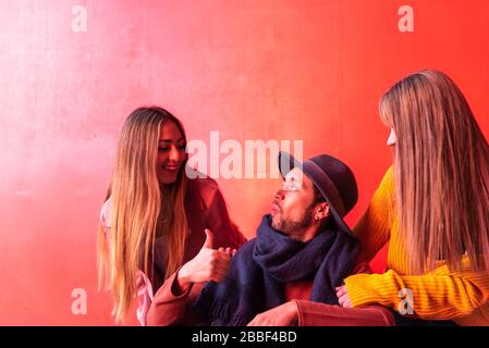 Lateinamerikanischer paraplegischer junger Mann, der glücklich neben zwei kaukasischen blonden jungen Mädchen mit rotem Hintergrund sitzt