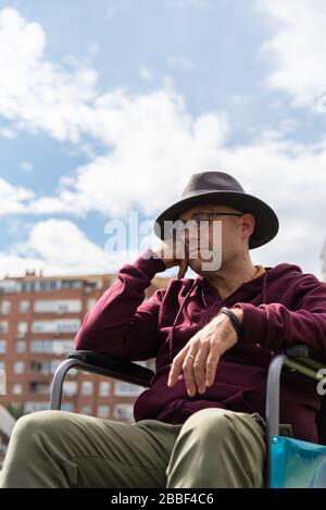 Kaukasischer Mann mit Brille und Hut depressiv und traurig in einem einsamen Rollstuhl in einem Park mit Himmel und einigen Gebäuden im Hintergrund mit Raum Stockfoto