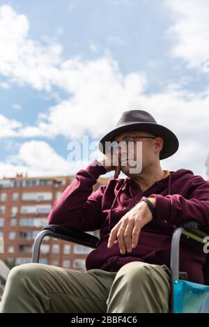 Kaukasischer Mann mit Brille und Hut depressiv und traurig in einem einsamen Rollstuhl in einem Park mit Himmel und einigen Gebäuden im Hintergrund mit Raum Stockfoto