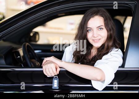 Attraktive junge Frau lacht glücklich in einem Auto in einem Autohaus und hält den Autoschlüssel Stockfoto