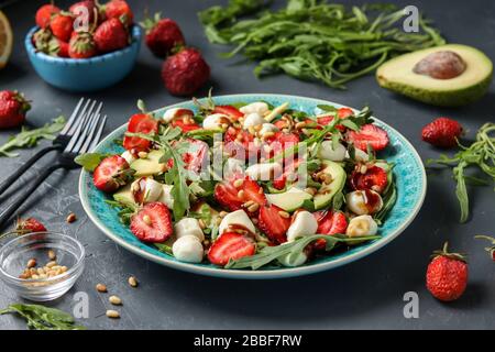 Gesunder Salat mit Erdbeeren, Avocado, Arugula und Mozzarella, gekleidet mit Olivenöl und Balsamico-Dressing in einem blauen Teller auf dunklem Hintergrund Stockfoto