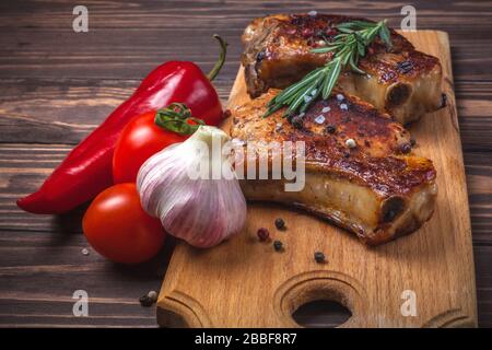 Gegrilltes Schweinesteaks und Gemüse auf einem rustikalen Holztisch. Gegrilltes Fleisch am Knochen mit Gewürzen, Knoblauch, Paprika, Tomaten und Rosmarin. Restaurant-Menü Stockfoto