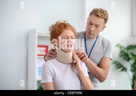 Männliche Physiotherapeutin untersucht Nackenfixator, weibliche Patientin vor Schmerzen erfroren Stockfoto