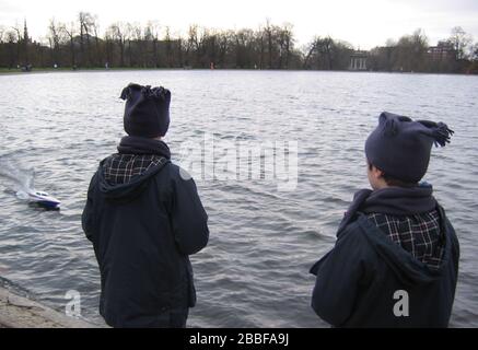 Winter Outdoor 2 Jungen tragen Hüte Mäntel und Schals mit Schnellboot mit Fernbedienung auf Kensington Gardens, Bootssee in London England GB Stockfoto