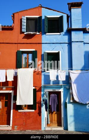 Venedig, Italy-Februar 2020; niedriger Blickwinkel auf einen Abschnitt einiger farbenfroher Häuser in Burano; eine blaue, eine rote, Kleidung, die an den Wäscheklammern hängt Stockfoto