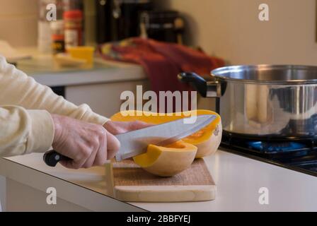 Frau, die Butternusskürbis auf Holzschneidebrett in der Küche neben vier Kochplatten für Butternusskürzersuppe schneidet Stockfoto