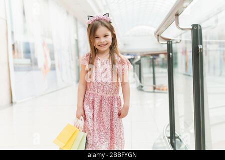 Porträt eines kleinen glücklichen Mädchens in der Mall. Ein lächelndes lachendes Mädchen in einem rosafarbenen Kleid mit einem niedlichen Rand mit Ohren und mit mehrfarbigen Taschen in ihren Händen läuft um das Einkaufszentrum und schaut auf die Kamera. Stockfoto