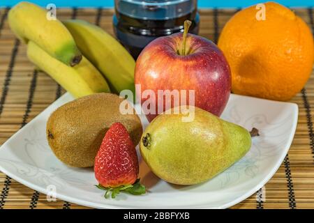Frische tropische und subtropische Früchte in einem großen weißen Gericht. Ein Glas Rotwein auf dem Wicker Bambus steht auf dem Esstisch. Nahaufnahme, selektiver Fokus. Stockfoto