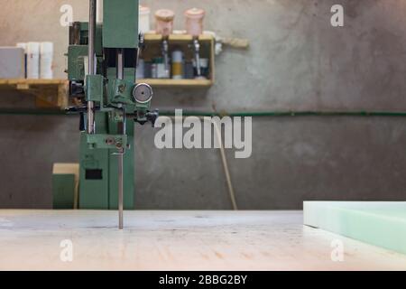 Prozess der Möbelherstellung. Ein junger Mann in einem dunklen Mantel Schnitt den Schaum für das Sofa auf einer Schneidemaschine. Er trägt eine Schutzbrille und Handschuhe. Stockfoto