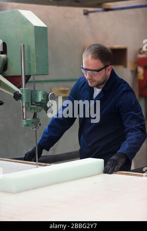 Prozess der Möbelherstellung. Ein junger Mann in einem dunklen Mantel Schnitt den Schaum für das Sofa auf einer Schneidemaschine. Er trägt eine Schutzbrille und Handschuhe. Stockfoto