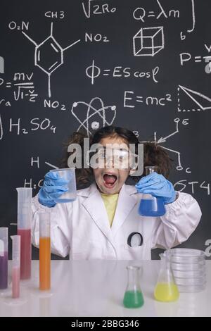 Lustige Wissenschaftlerin Mädchen mit Handschuhen und Brille im Labormantel mit Chemiekolben, Tafel Hintergrund mit wissenschaftlichen Formeln, Explosion im Laborgerät Stockfoto