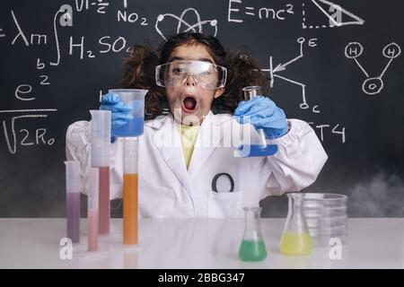 Überraschtes Mädchen der Wissenschaftlerin mit Handschuhen und Brille im Labormantel mit chemischen Kolben, Hintergrund auf der Tafel mit wissenschaftlichen Formeln, Explosion in der Arbeit Stockfoto