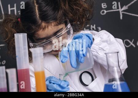 Kleines Wissenschaftlermädchen mit Brille und Handschuhen im Labormantel, das chemische Flüssigkeiten in Kolben mischt, Hintergrund auf der Tafel mit wissenschaftlichen Formeln, Explosion in Stockfoto