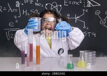 Lustige Wissenschaftlerin Mädchen mit Handschuhen und Brille im Labormantel mit Chemiekolben, Tafel Hintergrund mit wissenschaftlichen Formeln, Explosion im Laborgerät Stockfoto