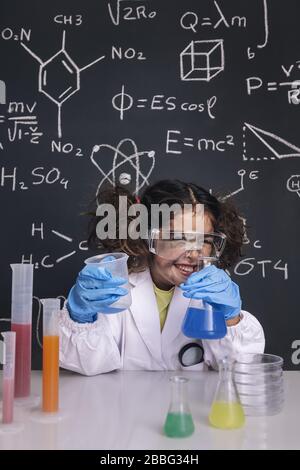 Lustige Wissenschaftlerin Mädchen mit Handschuhen und Brille im Labormantel lachen, Hintergrund auf der Tafel mit wissenschaftlichen Formeln, Explosion im Labor, zurück zu s Stockfoto