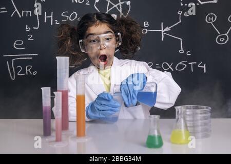 Lustige Wissenschaftlerin Mädchen mit Handschuhen und Brille im Laborkittel, die chemische Kolben mischt, Hintergrund auf der Tafel mit wissenschaftlichen Formeln, Explosion im Labor Stockfoto
