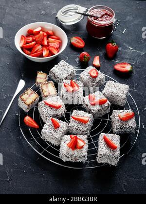 Klassische australische Lamingtons Vanilleschwamm Minikuchen mit Erdbeermarmeladenschicht im Inneren, oben mit Schokolade und Kokos auf einem dunklen Beton beschichtet Stockfoto