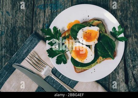 Toast mit Avocado, gekochtem Eierspinat und Petersilie auf weißem Teller mit Messer und Gabel auf Serviette und rustikalem blauen Holzhintergrund. Stockfoto