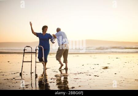 Ein älterer Mann und eine Krankenschwester tanzten zusammen neben einer Gehhilfe am Strand. Stockfoto