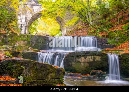 Sonniger Tag im Sommerwald. Bogen einer alten Steinbrücke. Kleiner Fluss und mehrere natürliche Wasserfälle Stockfoto