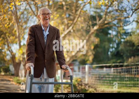 Lachender älterer Mann, der Spaß hat, durch einen Park mit einem Wanderrahmen zu gehen. Stockfoto