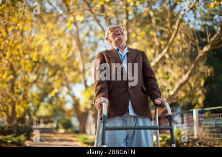Lachender älterer Mann, der Spaß hat, durch einen Park mit einem Wanderrahmen zu gehen. Stockfoto
