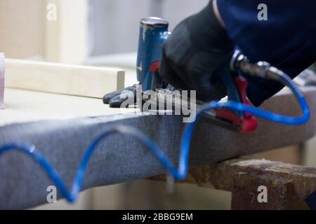 Prozess der Möbelherstellung. Ein junger Mann in einem dunklen Mantel, der einen Teil des Sofas mit einem Hefter auf einem Schreibtisch zusammenstellt. Er hat eine schwarze sa Stockfoto