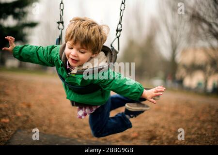 Fröhlicher kleiner Junge, der auf einem Kettenschwung in einem Park spielt. Stockfoto