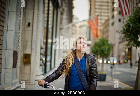 Fröhliche junge Frau, die ihr Fahrrad auf einem Bürgersteig neben einer städtischen Straße spazieren ging Stockfoto