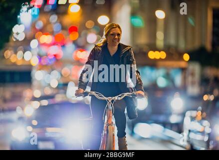 Lächelnde junge Frau, die nachts mit dem Fahrrad auf einer belebten Stadtstraße unterwegs ist Stockfoto