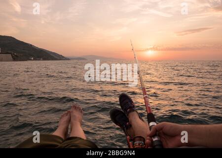 Es gibt ein Paar und sie angeln, während die Sonne untergeht. Sie sitzen auf einem Gerüst und einer von ihnen hält Angelrute gegen Sonnenuntergang. Stockfoto