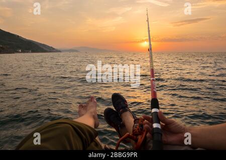 Es gibt ein Paar und sie angeln, während die Sonne untergeht. Sie sitzen auf einem Gerüst und einer von ihnen hält Angelrute gegen Sonnenuntergang. Stockfoto