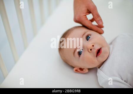 Niedlicher Babyjunge, der aus seiner Krippe hochschaut, während seine junge Mutter sanft ihre Hand auf seine Wange legt Stockfoto