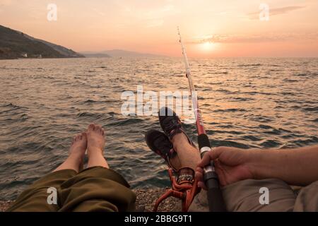 Es gibt ein Paar und sie angeln, während die Sonne untergeht. Sie sitzen auf einem Gerüst und einer von ihnen hält Angelrute gegen Sonnenuntergang. Stockfoto