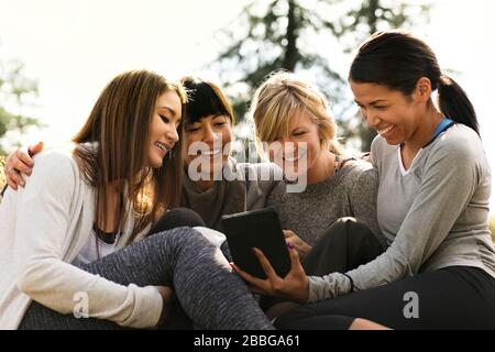 Vier glückliche Freundinnen, die gemeinsam in einem sonnigen Park ein digitales Tablet betrachten Stockfoto