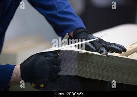 Prozess der Möbelherstellung. Ein junger Mann in einem dunklen Mantel, der die überschüssigen Teile des Sofas auf einem Schreibtisch ausgeschnitten hat. Stockfoto