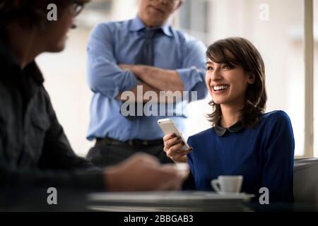 Lächelnde junge Geschäftsfrau, die ein Handy in einem Treffen mit zwei Kollegen hält Stockfoto